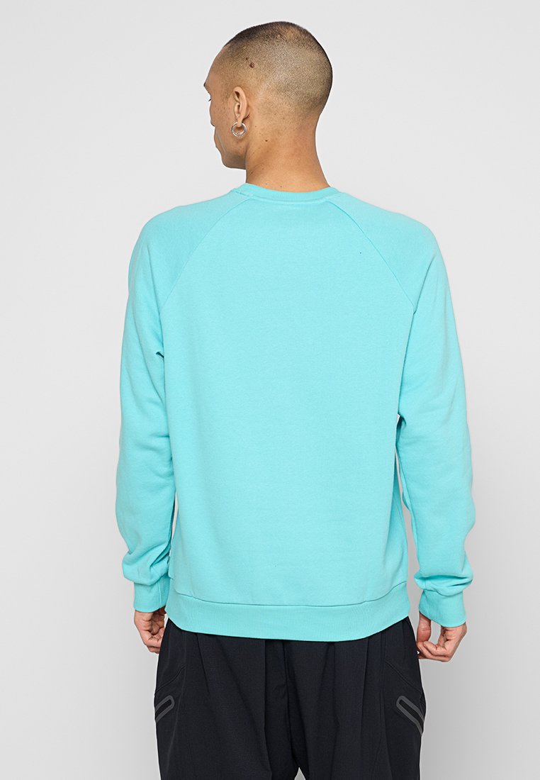Joven con cabeza rapada y pendientes de aro, lleva una sudadera de manga larga azul claro y pantalones negros, de espaldas a la cámara.