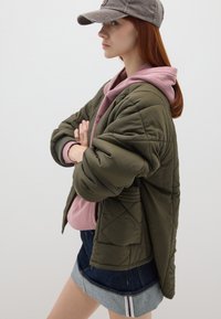 Giacca trapuntata verde oliva sopra una felpa con cappuccio rosa, abbinata a una gonna di jeans. Il cappellino è grigio con un logo frontale.