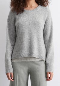 Pull gris tricoté avec un col rond côtelé, épaules tombantes et manches longues. Texture duveteuse et coupe décontractée, superposé à un haut gris clair.