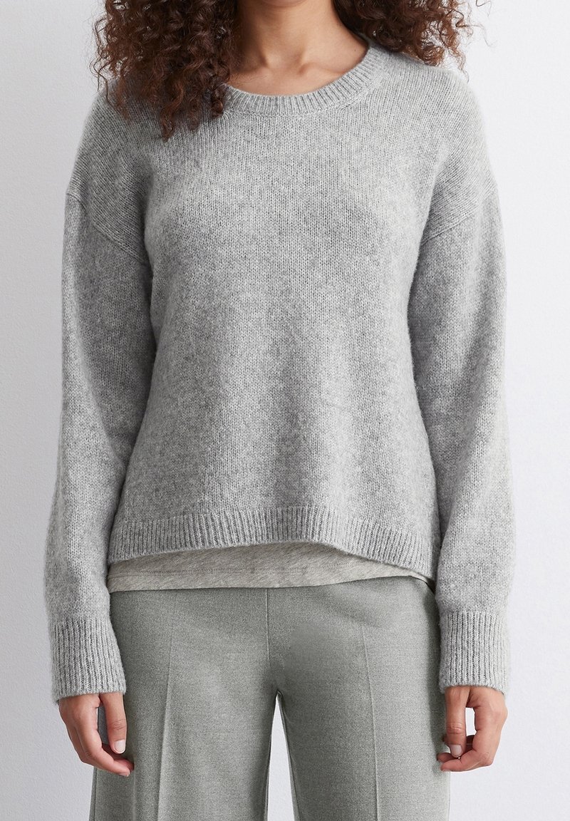 Pull gris tricoté avec un col rond côtelé, épaules tombantes et manches longues. Texture duveteuse et coupe décontractée, superposé à un haut gris clair.