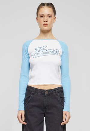 White cropped shirt with light blue long sleeves. Features a blue graphic text "Kanz est. 1989" on the chest. Black high-waisted pants visible.