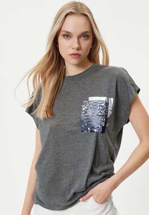 Grå kortærmet t-shirt med en skinnende pailletlomme i forskellige nuancer af blå og sølv. Har en afslappet pasform og blød tekstur.
