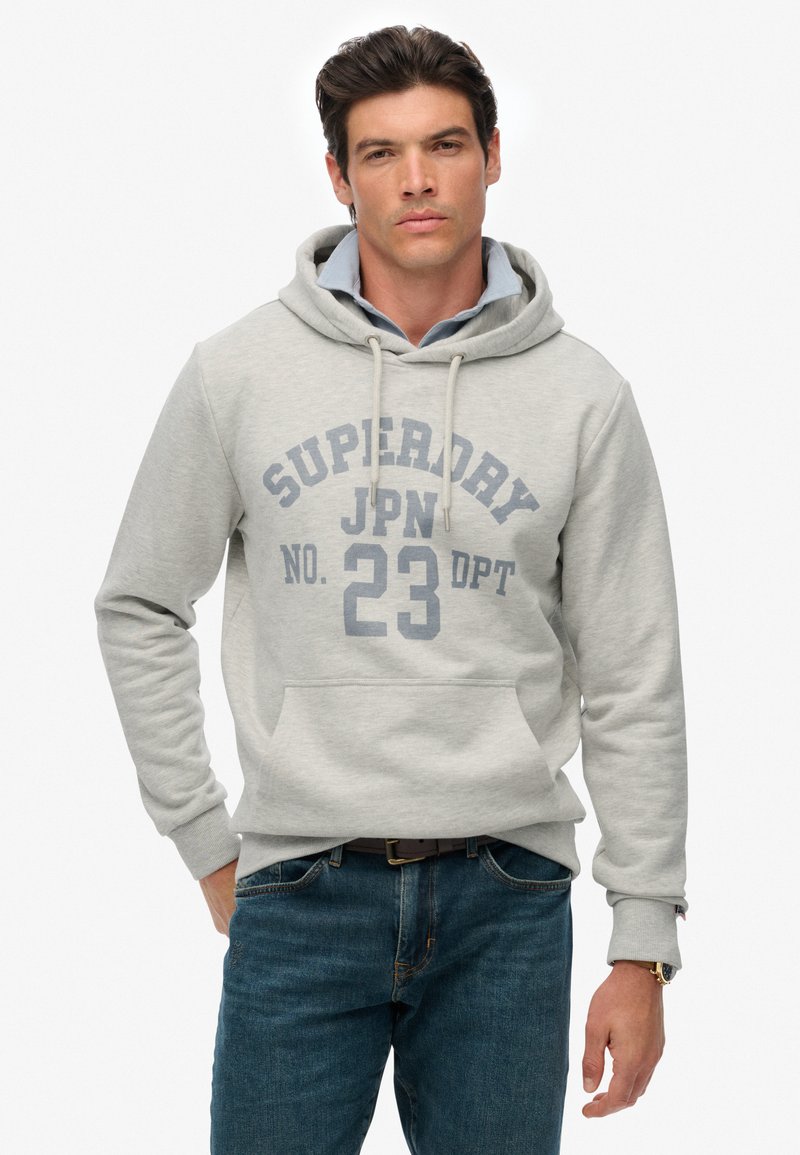 Man in een lichtgrijze Superdry hoodie met trekkoorden over een blauwe colbert en blauwe jeans, staande tegen een witte achtergrond.