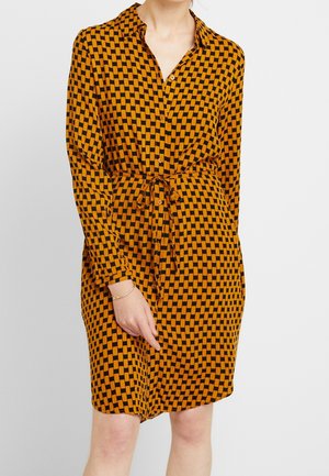 Mujer con un vestido de botones de manga larga con un patrón de cuadros naranja y negro y un cinturón anudado en la cintura.