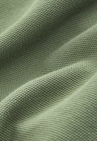 Lichtgroene gestructureerde stof met een fijn gebreid patroon, met zachte plooien en subtiele schaduwen die de textuur van het materiaal benadrukken.