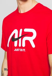 Punainen puuvillainen t-paita, jossa on näkyvä valkoinen "AIR"-logo ja musta Nike swoosh -merkki, sekä teksti "JUST DO IT." alla. Klassinen pyöreä kaulus.