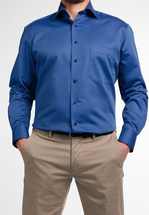Mężczyzna w niebieskiej, długorękawowej koszuli zapinanej na guziki z kieszenią na piersi oraz beżowych spodniach, z rękami w kieszeniach.