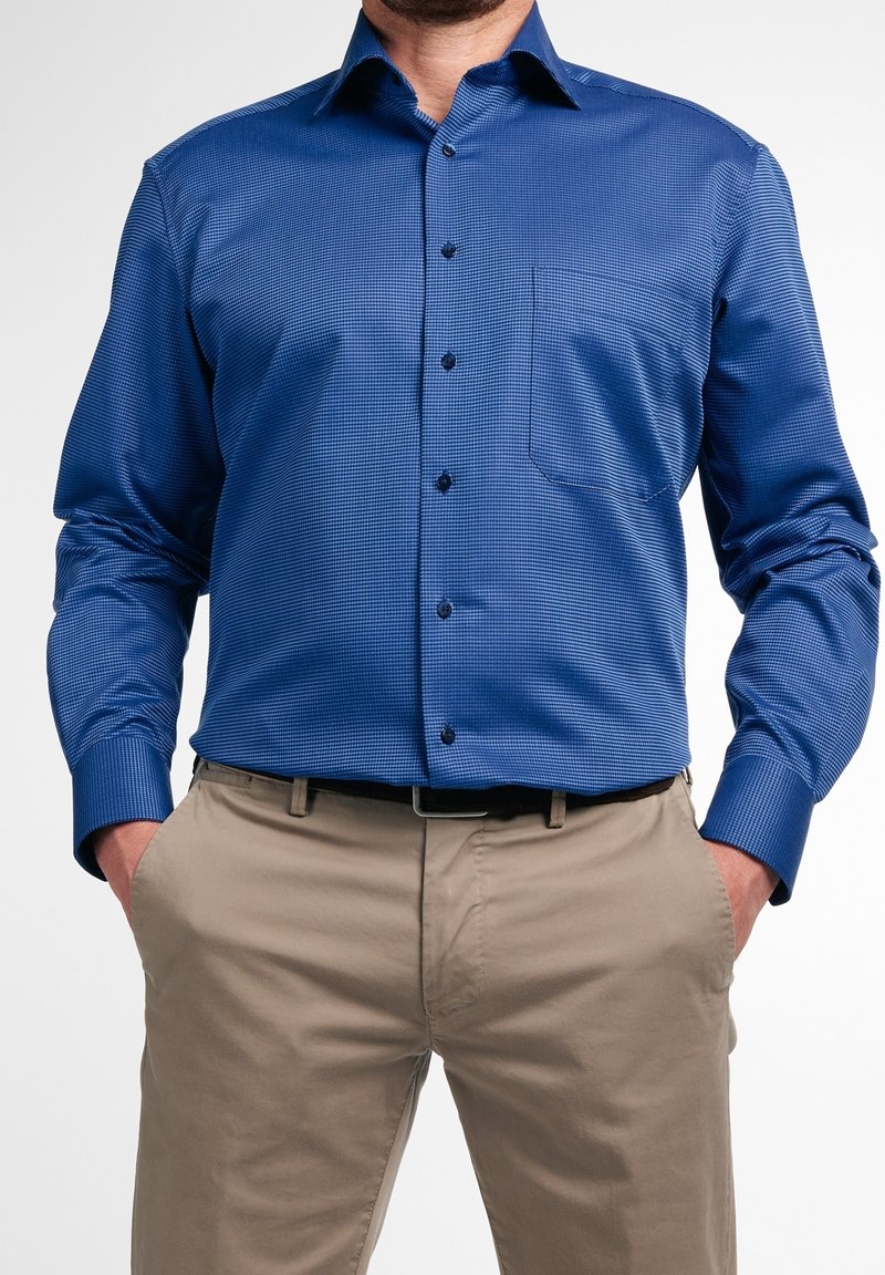 Homme portant une chemise bleue à manches longues avec des boutons sur le devant et une poche poitrine, ainsi qu'un pantalon beige, les mains dans les poches.