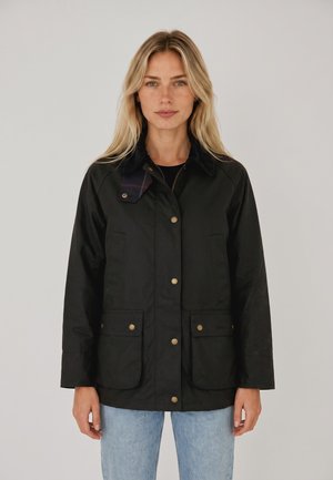 Femme blonde aux cheveux lisses portant une veste noire boutonnée avec des poches avant et un jean bleu clair sur un fond uni.