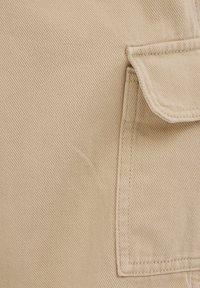 Bežs audums ar teksturētu ribbota modeli, kas satur taisnstūra kabatu. Malas ir šūtas, uzsverot izturību.