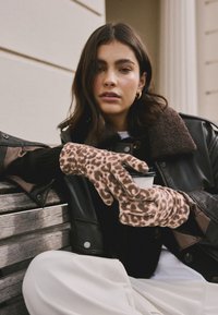 Bruine handschoenen met luipaardprint gemaakt van zacht materiaal, met een aangesloten ontwerp. Een persoon houdt een kopje vast terwijl hij een zwarte leren jas draagt.