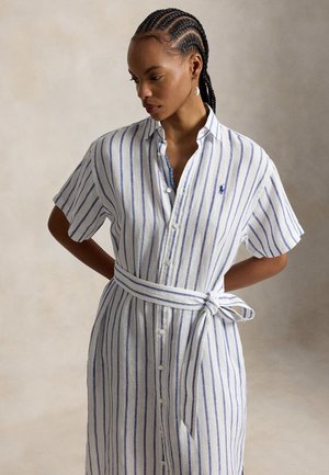 Frau mit geflochtenem Haar, die ein weißes Kleid mit blauen vertikalen Streifen und einem gebundenen Gürtel trägt, steht mit den Händen hinter dem Rücken.