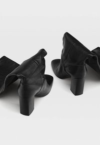 Bottes en cuir noir mi-mollet avec des talons hauts épais, des pointes pointues et un design décontracté sur un fond léger uni.