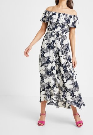 Kvinde iført sort-hvid blomsteret off-shoulder midi-kjole med flæsedetalje, bundet talje og lyserøde åbne tå-sandaler, stående på en ensfarvet baggrund.