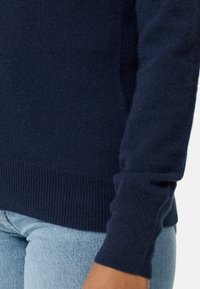 Dunkelblauer gestrickter Pullover mit gerippten Bündchen und Saum. Weiche Textur mit enger Passform, teilweise über hellblauen Jeans sichtbar.