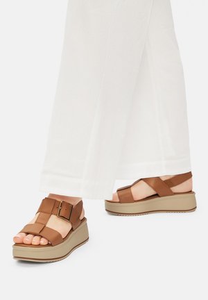 Füße in weißen Hosen mit weitem Bein, die braune Plateau-Sandalen mit Schnallenriemen auf einer hellbeigen Sohle tragen, stehen auf einer weißen Oberfläche.