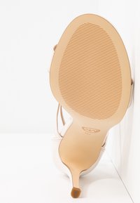 Sandale à talon haut beige pour femme, inclinée pour montrer la semelle avec un motif de bande de roulement texturé et un talon aiguille fin sur une surface blanche.