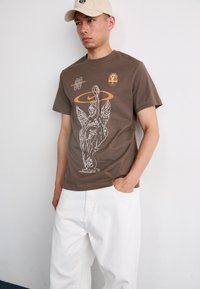 Brun bomull T-skjorte med et grafisk design som viser en statue med vinger og en oransje Nike-logo, iført hvite bukser.