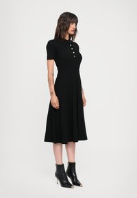 Black ribbed dress with short sleeves, button detail at the collar, and pleated skirt. Worn with black ankle boots featuring gold accents.