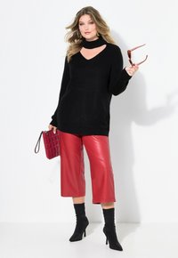 Schwarzer Strickpullover mit V-Ausschnitt und Ausschnitten, kombiniert mit roten Kunstleder-Culottes und schwarzen Ankle Boots. Hält rote Sonnenbrille und Tasche.