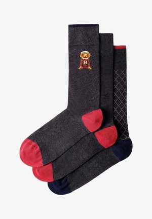Tres pares de calcetines gris oscuro con dedos y talones de colores; uno tiene un perro con un jersey de rayas, otro es liso, el tercero tiene un patrón de diamantes.