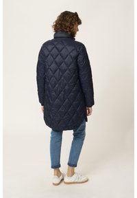 Abrigo largo acolchado de color azul marino con cuello alto, con un patrón de diamantes, combinado con jeans de color azul claro y zapatillas blancas.