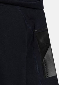 Felpa navy in tessuto testurizzato, con un patch in gomma nera sulla manica e cuciture in contrasto lungo le cuciture.