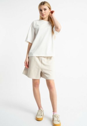 Młoda kobieta stojąca w beżowych, casualowych szortach, białym oversize t-shircie i beżowych trampkach z różowymi skarpetkami na białym tle.