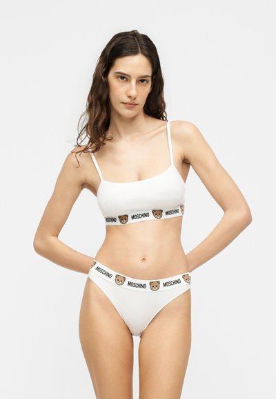 Femme portant un bralette blanc Moschino et un slip assorti avec des bandes au logo ours en peluche, debout, les mains derrière le dos.