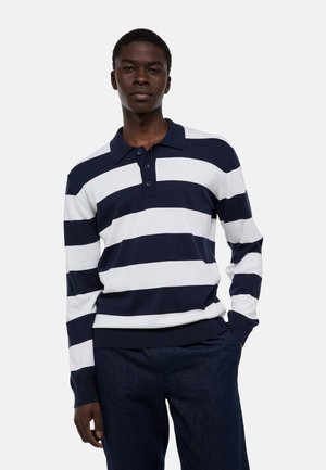 Jeune homme portant un polo à manches longues à larges rayures bleu marine et blanc, et un pantalon foncé, debout avec une main dans la poche.