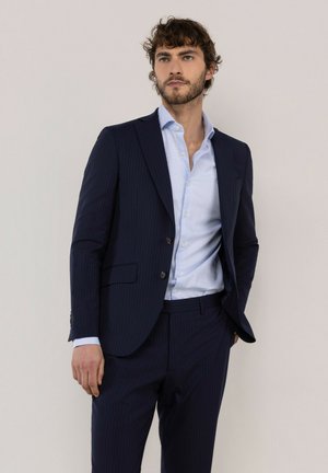 Hombre con cabello rizado y barba, vestido con un traje azul marino de rayas finas y una camisa azul clara, de pie con una mano en el bolsillo.