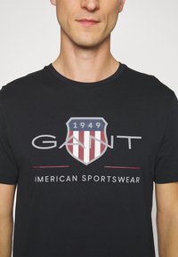 Svart bomull T-skjorte med et skjoldlogo med røde og hvite striper, preget tekst "GANT AMERICAN SPORTSWEAR" under. Rund utkledning.