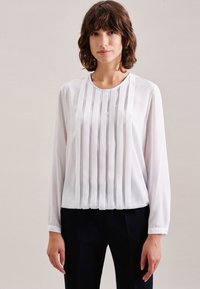 Blusa bianca a maniche lunghe con pieghe verticali e scollatura rotonda, realizzata in tessuto morbido. Abbinata a pantaloni scuri su misura.