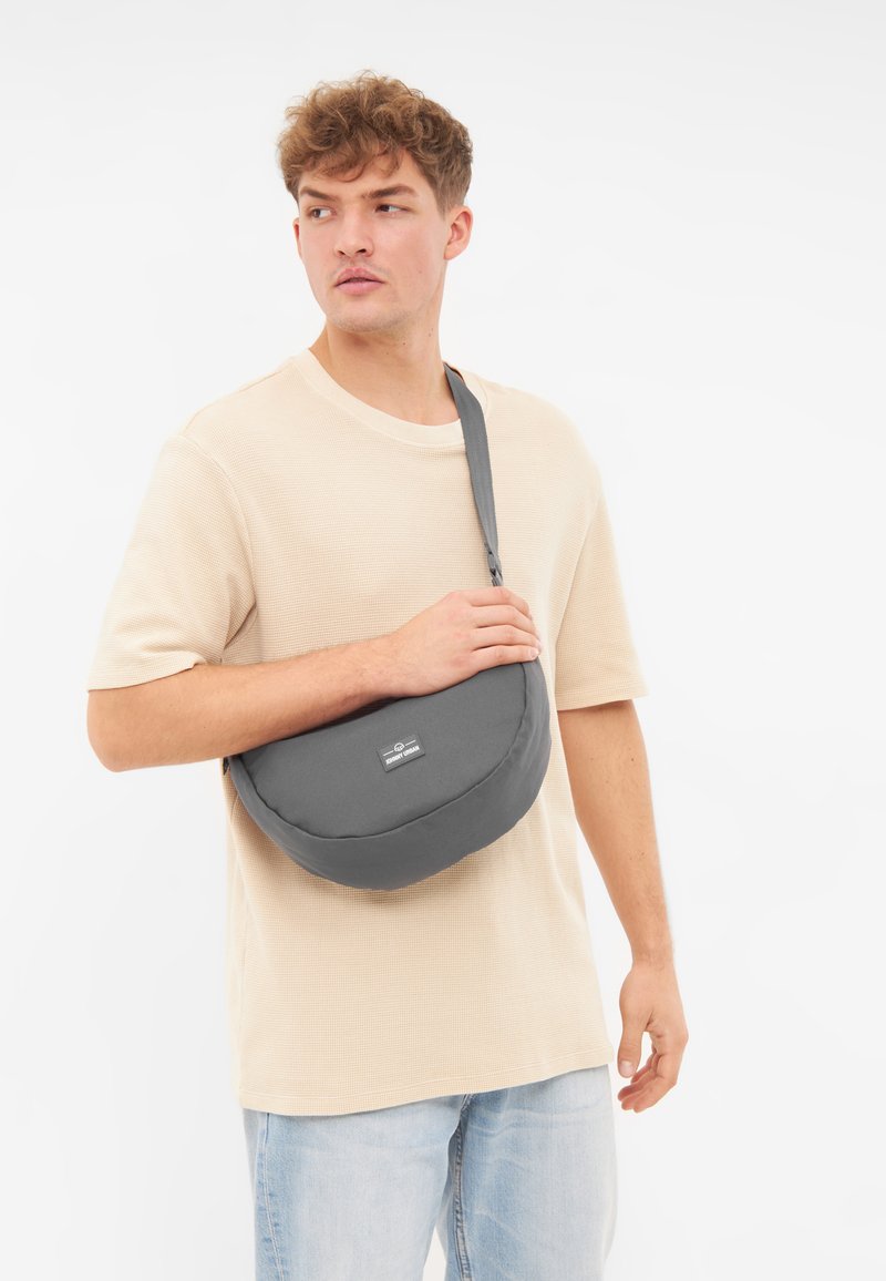 Borsa a spalla grigia, realizzata in tessuto morbido, di forma arrotondata con un fronte piatto, tracolla regolabile e una piccola etichetta con logo sul davanti.