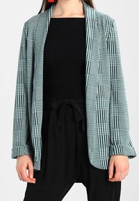 Rutig blazer i teal och svart, tillverkad av lättviktigt material med avslappnad passform, sjalkrage och uppvikta ärmslut. Bärs över en svart topp.