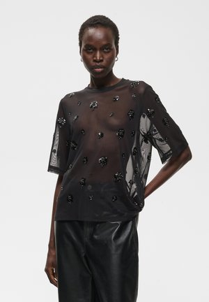 Femme à la peau foncée porte un haut noir transparent à manches courtes avec des ornements floraux noirs et un pantalon en cuir noir, posant avec une main sur la hanche.