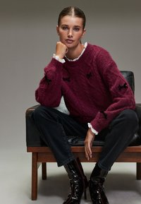 Weinroter Strickpullover mit strukturiertem Muster und schwarzen Schleifenakzenten, kombiniert mit dunklen Jeans und glänzenden schwarzen Stiefeletten.