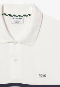 Polo blanco con cuello de canalé, que presenta detalles en verde y negro, dos botones y un pequeño logo bordado de un cocodrilo en el pecho.