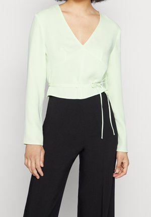 Femme portant un haut cache-cœur vert clair à manches longues avec un lien sur le côté et un pantalon noir taille haute à jambes larges sur un fond uni.