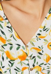 Kukallinen mekko, jossa on valkoinen pohja, kelta-oransseja sitruskuvioita ja vihreitä lehtiä; V-pääntie ja kultaiset napit tuovat yksityiskohtia.