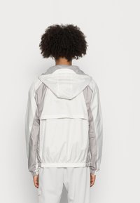 Vit och ljusgrå vindjacka med huva, elastiska ärmslut och en mesh-fodrad huva, med en horisontell bakficka.