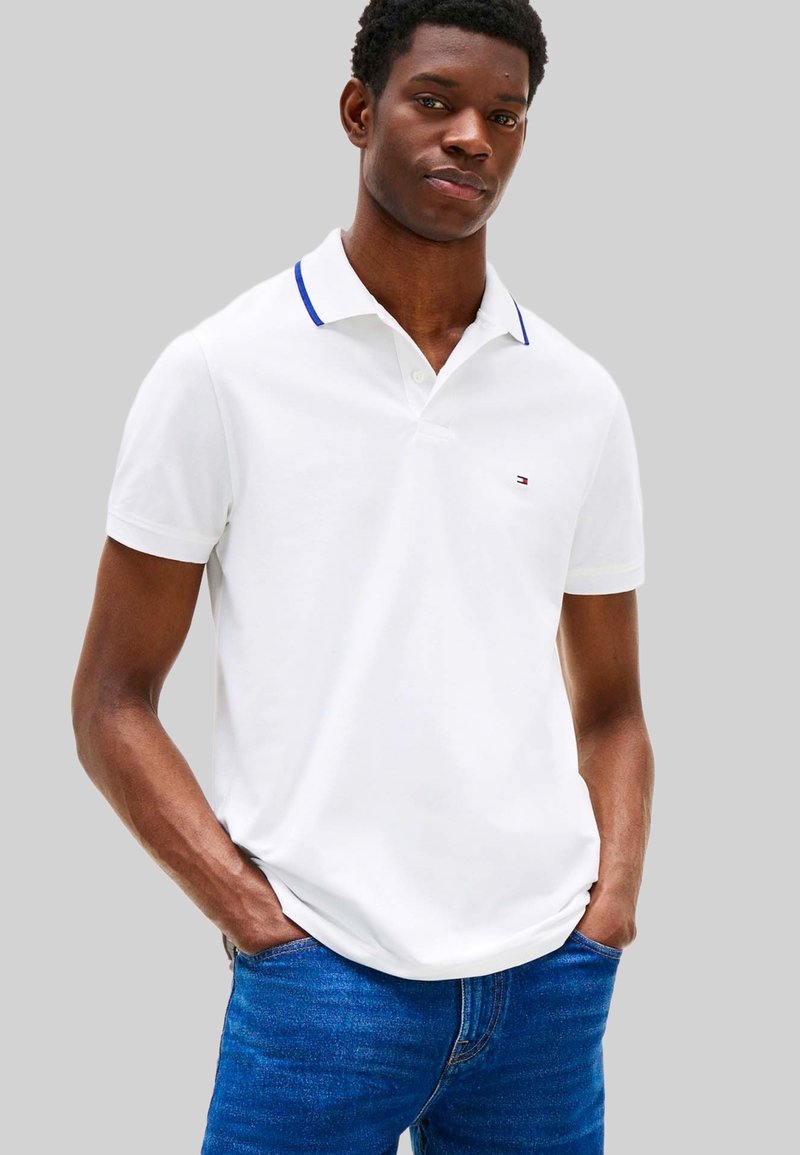 Wit poloshirt met blauwe accenten op de kraag, korte mouwen en een logo op de borst, gecombineerd met een blauwe spijkerbroek. Effen achtergrond.