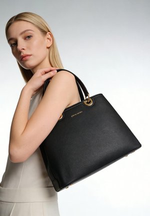 Femme blonde portant une robe blanche sans manches tient un grand sac à main noir Pierre Cardin sur l'épaule devant un fond uni.