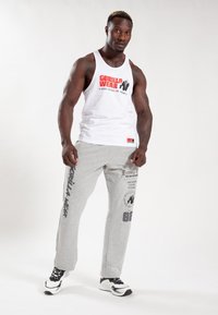 Witte tanktop met rood gorilla-logo en tekst "GORILLA WEAR", gecombineerd met grijze sweatpants met merknaam en graphics. Zwart-witte sneakers.