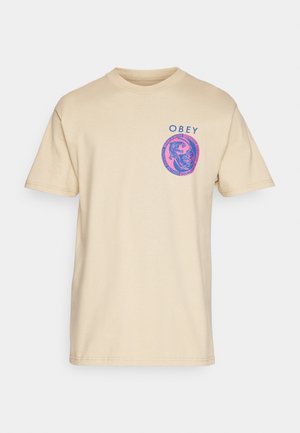 Camiseta de algodón beige con mangas cortas y cuello redondo. La impresión gráfica presenta el texto azul "OBEY" y un diseño circular rosa en el área izquierda del pecho.