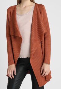Rust-colored cardigan with a draped front, ribbed texture, and diagonal stripes. Worn over a light pink fitted top and black pants.