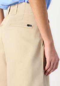 Pantalons beige avec une texture lisse, dotés de poches arrière et d'une petite étiquette logo. La vue en gros plan montre la ceinture et les détails de la couture latérale.