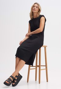 Zwarte geribbelde midi-jurk met korte mouwen, zijsplit en zwarte platform sandalen op een houten kruk tegen een witte achtergrond.