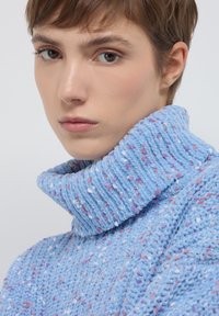 Maglione a maglia blu chiaro con colletto alto in rilievo. Il tessuto testurizzato presenta spruzzi di rosa e bianco in tutto.