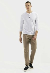 Langärmliges weißes Henley-Shirt mit drei schwarzen Knöpfen, kombiniert mit beigen Chinos und marineblauen Sneakers, vor einem schlichten Hintergrund stehend.